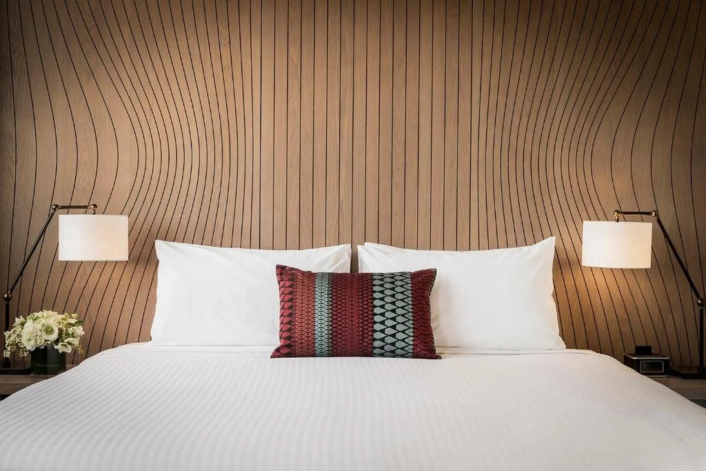 A neatly made bed with white linens, decorative pillow in front, two wall-mounted lamps on either side, a small plant with white flowers on the left, and a wooden slat wall background.