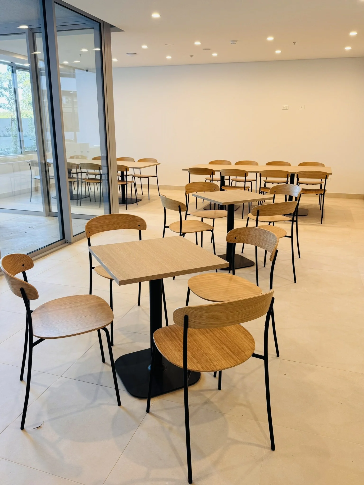 Empty modern cafe or dining area with wooden tables and chairs, large windows, and neutral-colored walls.