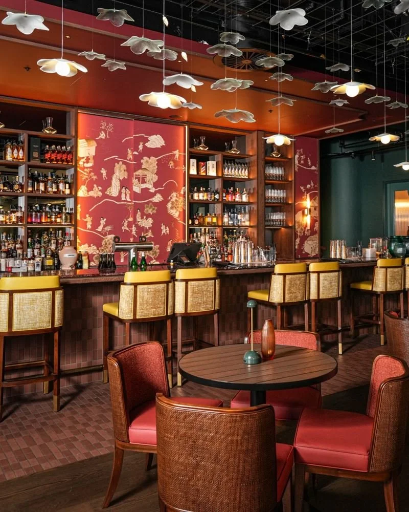 Interior of a bar with a decorated ceiling featuring cloud-shaped light fixtures, wooden shelves with liquor bottles, a red painted wall with gold Asian-inspired art, and seating including yellow-backed bar stools and red upholstered chairs around tables.