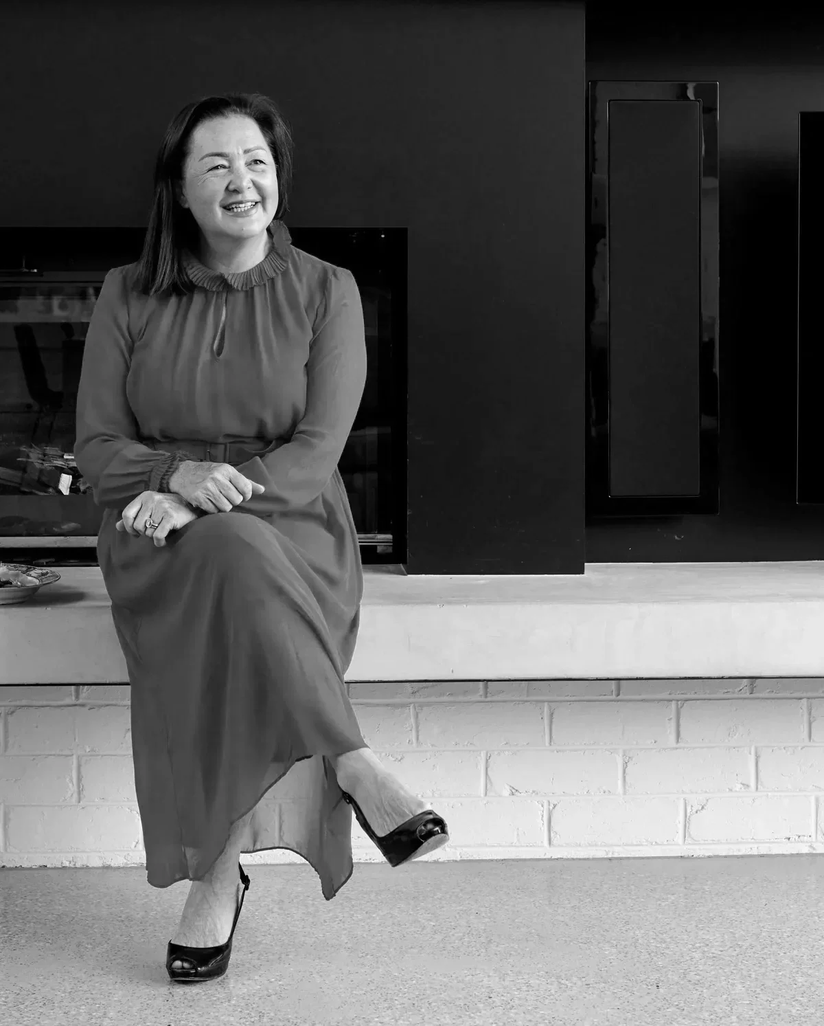 A woman with shoulder-length dark hair smiling, sitting on a fireplace hearth, wearing a long dress and high heels, in a black and white photo.