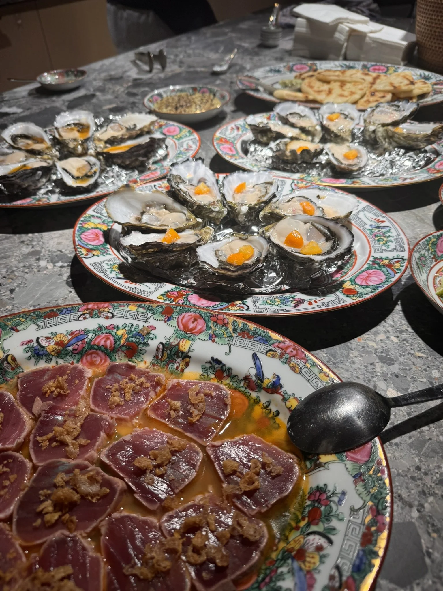 A table with plates of raw oysters on the half shell, topped with orange roe, and slices of raw meat garnished with condiments, along with cutlery and napkins.