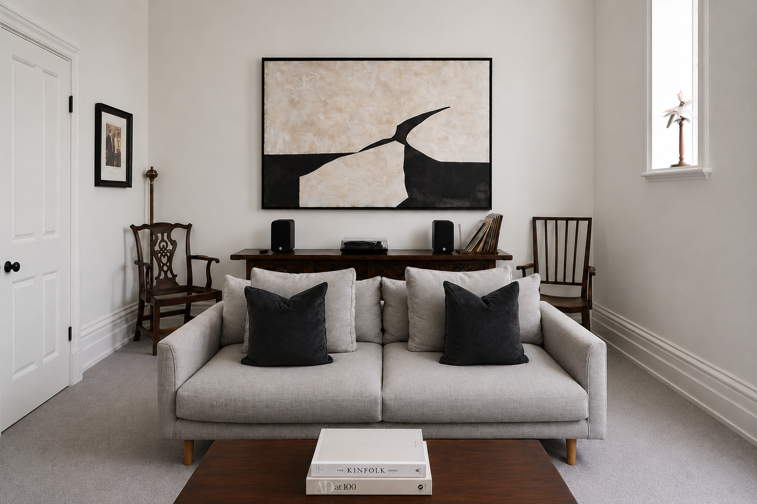 Living room with a gray sofa, black and gray pillows, wooden chairs, a wooden console with speakers and records, large abstract wall art, and a window with a plant.