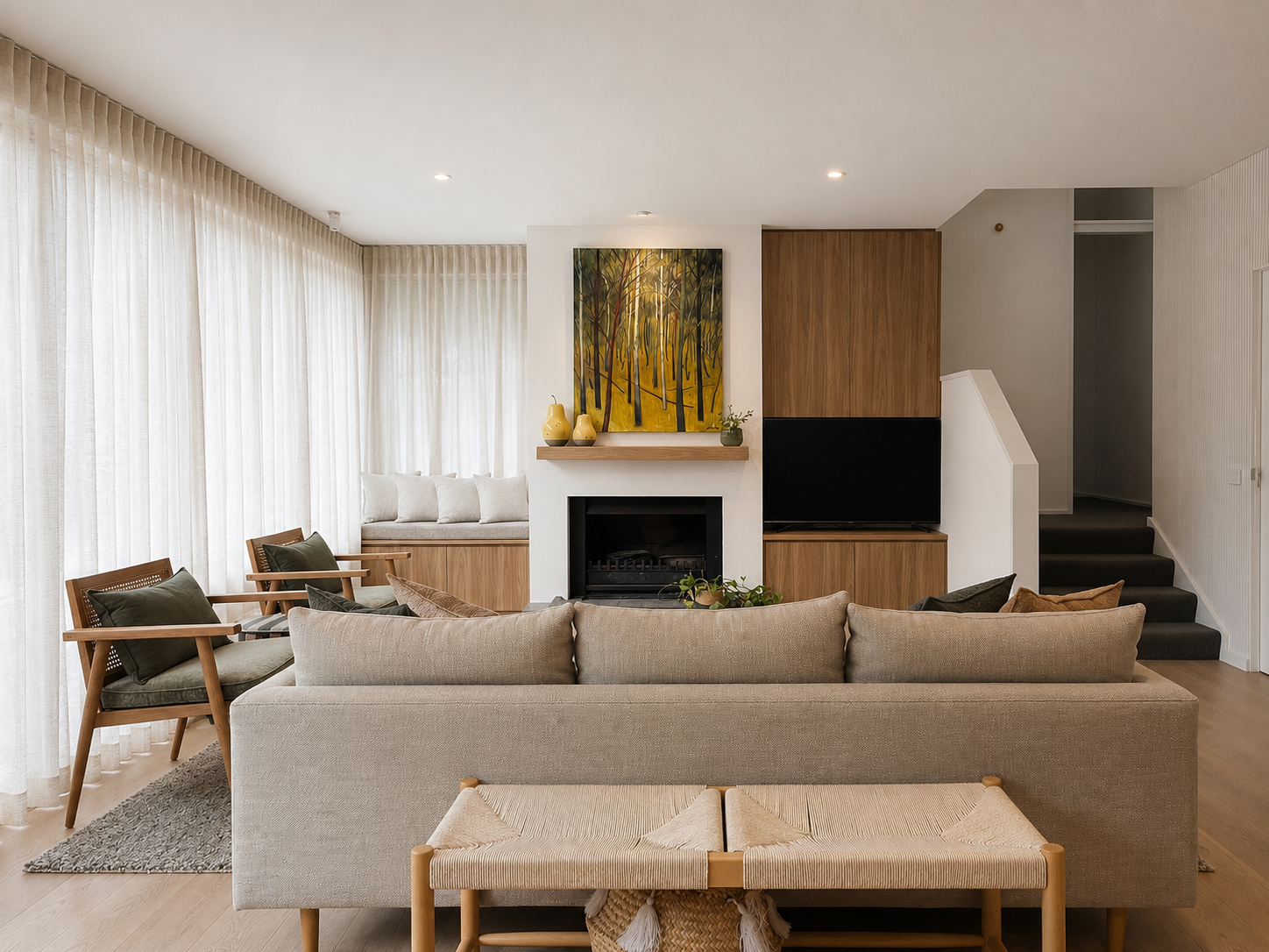 Living room with beige sofa, wooden armchairs with dark cushions, window seat with pillows, fireplace, wall-mounted TV, abstract forest painting, decorative vases, potted plant, and stairs.