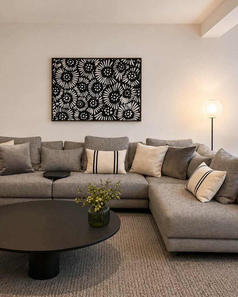 Living room with a grey sectional sofa, decorative pillows, a large black and white floral artwork on the wall, a black round coffee table with a small green plant, and a white floor lamp with a globe shade.
