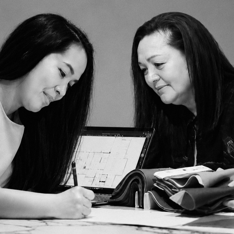 Two women discussing design plans at a table with fabric swatches and a laptop, with a decorative flower arrangement in the background.