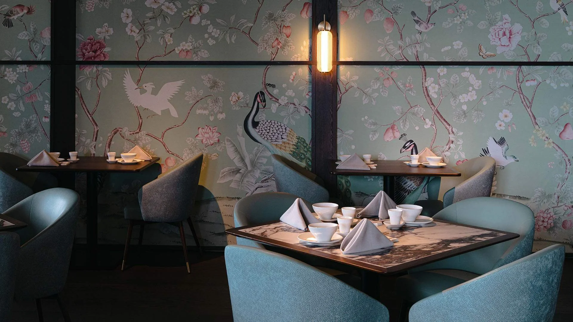 Dining area with tables set with white napkins, bowls, cups, and saucers, featuring patterned marble tabletops, pastel chairs, and a mural of peacocks, birds, and cherry blossoms on the wall, lit by a hanging lantern