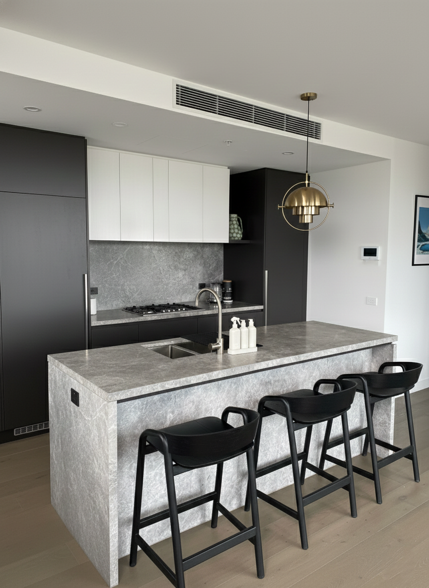Modern kitchen with gray stone island, black chairs, white and black cabinets, stainless steel sink, gold pendant light, and minimal decor.