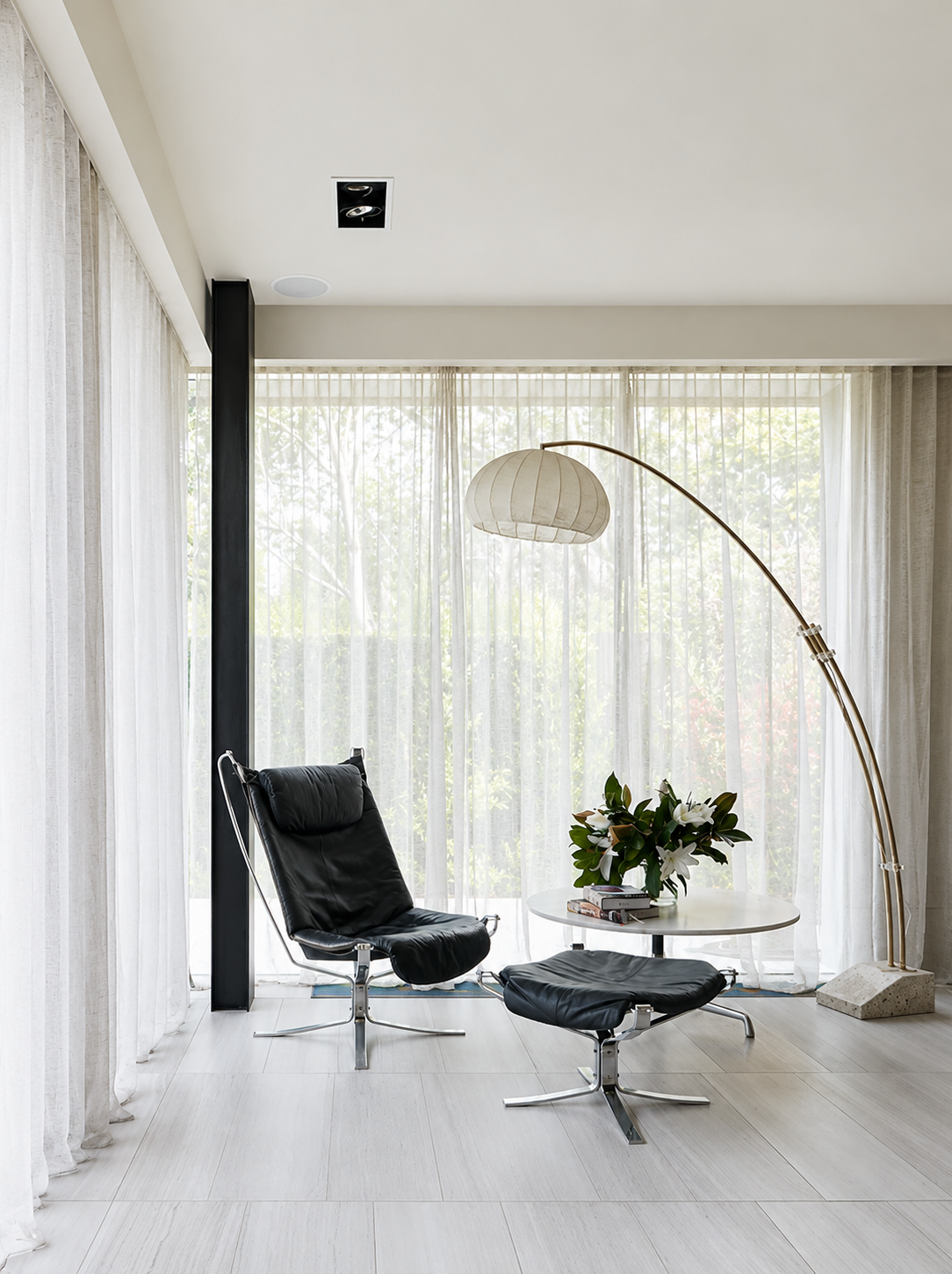 Modern living room with a black leather lounge chair and matching ottoman, a white round side table with books and a plant, a tall arc floor lamp, sheer white curtains, and large windows showing trees outside.