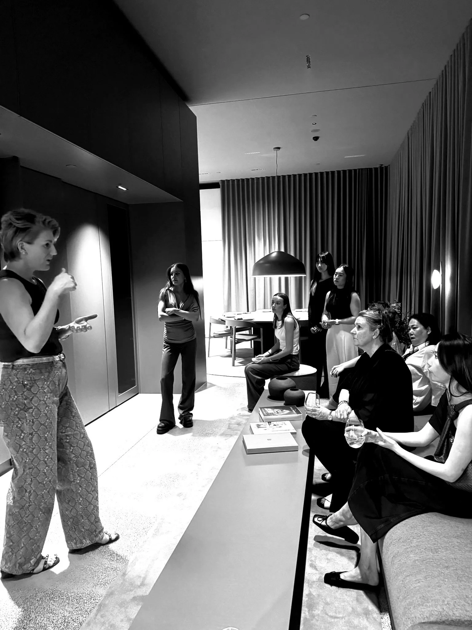 Group of women in a modern, minimalist room with curtains, sitting and standing, listening to a woman speaking.