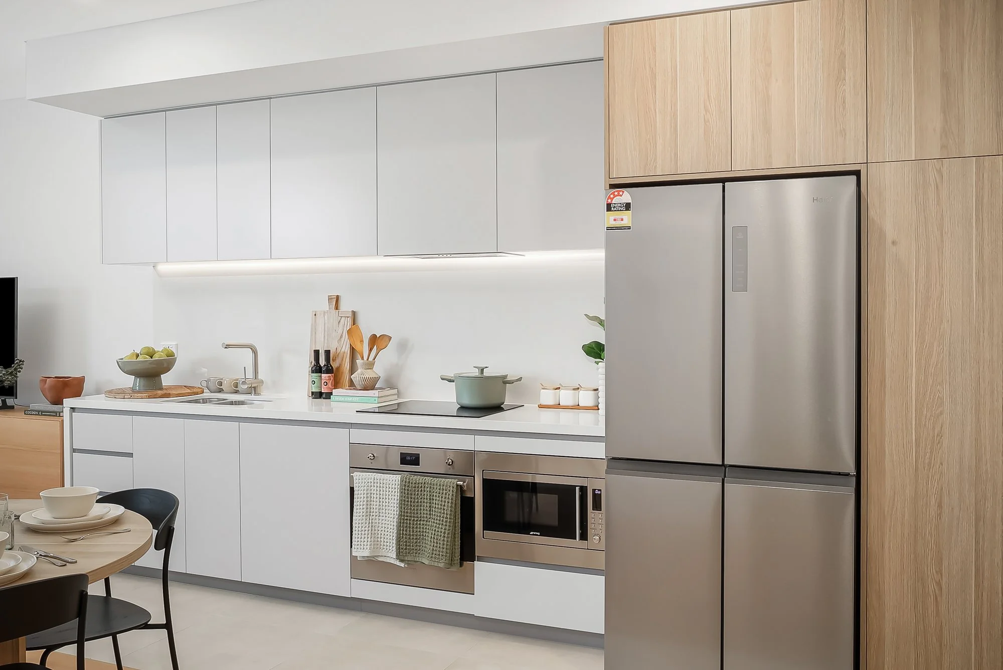 Modern kitchen with white and wood cabinetry, a stainless steel refrigerator, oven, microwave, and a small dining table with black chairs.