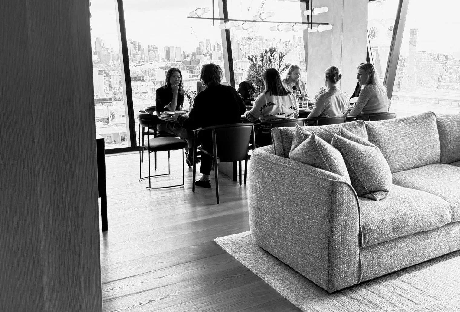 A group of six women sitting around a dining table in a bright, modern apartment with large windows showing a cityscape. The apartment has a sofa and a contemporary chandelier.