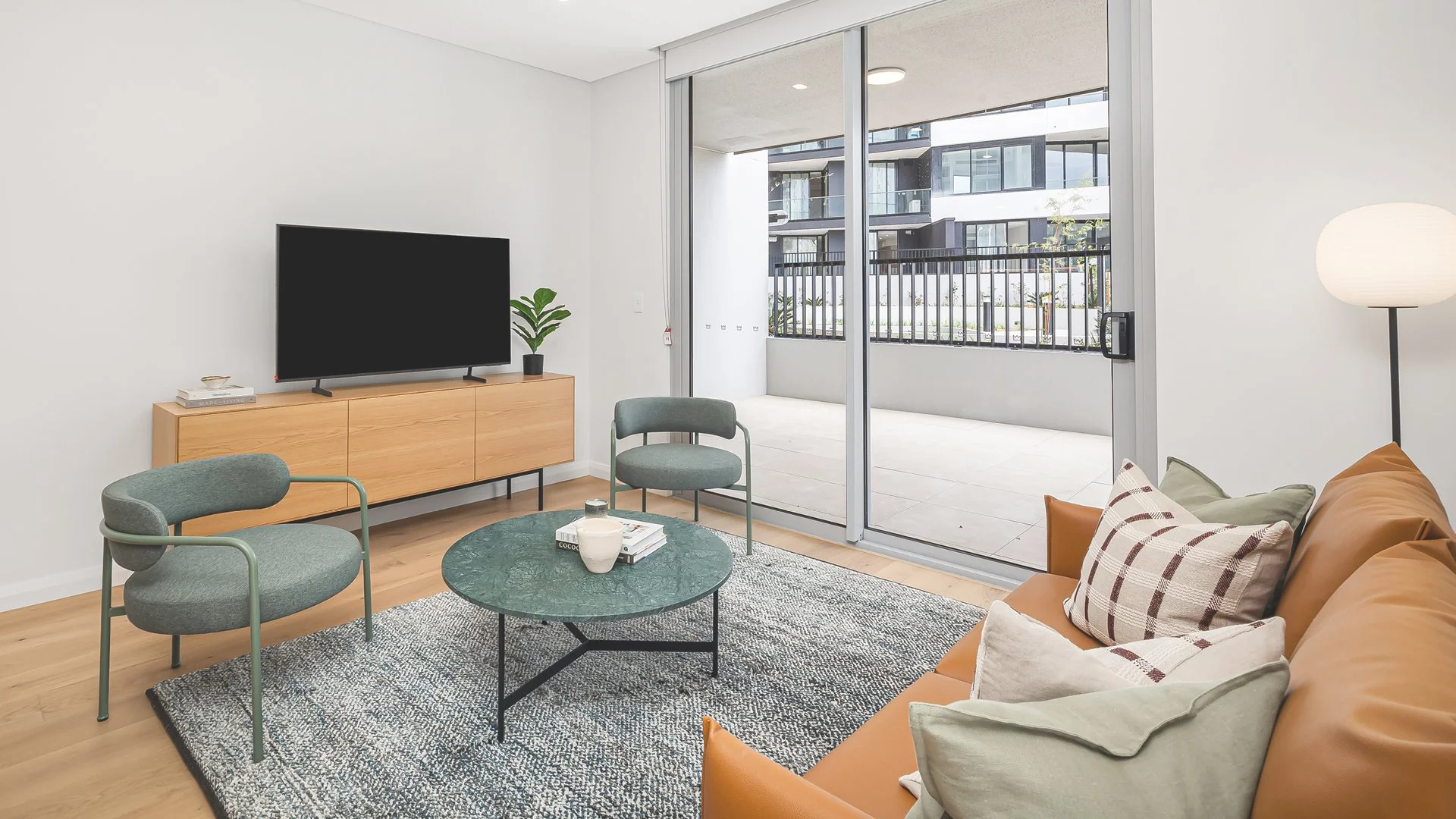 Living room with a brown leather sofa, green chairs, a round coffee table, a TV on a wooden console, and sliding glass door leading to a balcony with apartment buildings outside.