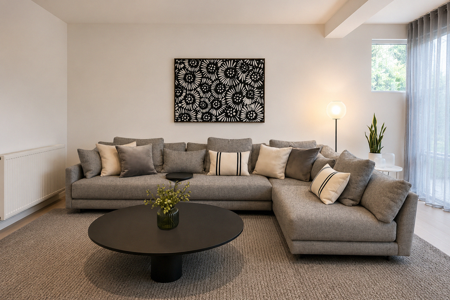 Modern living room with a gray sectional sofa, black round coffee table with green vase, artwork on the wall, potted plant, floor lamp, sheer curtains, and a window