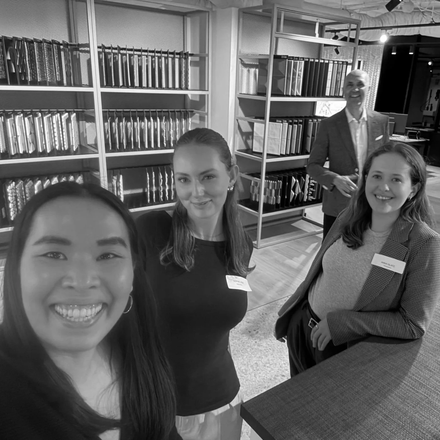 Four women and one man taking a selfie in an indoor setting with shelves of documents or binders in the background. The women are smiling and the man is standing behind them, also smiling. Some women are wearing name tags.