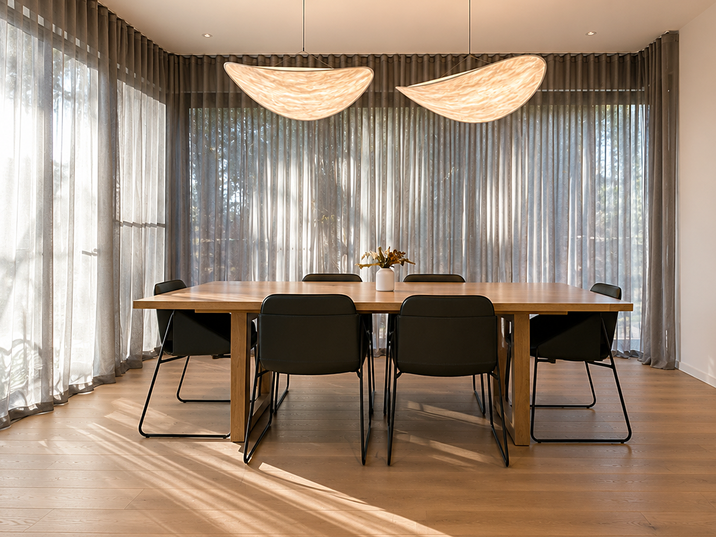Modern dining room with a wooden table, six black chairs, a white vase with flowers, large windows with sheer curtains, and two pendant light fixtures.