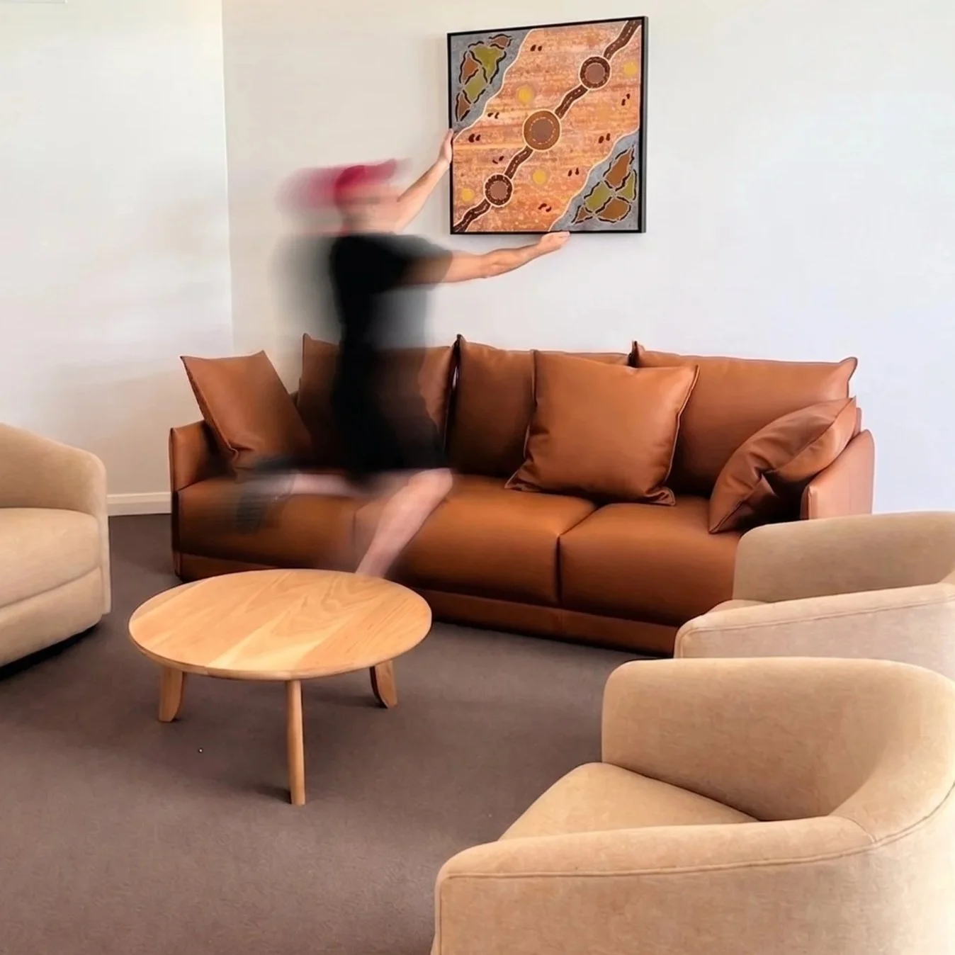 A person jumping on a brown leather sofa in a living room, with beige armchairs, a wooden coffee table, and abstract wall art.