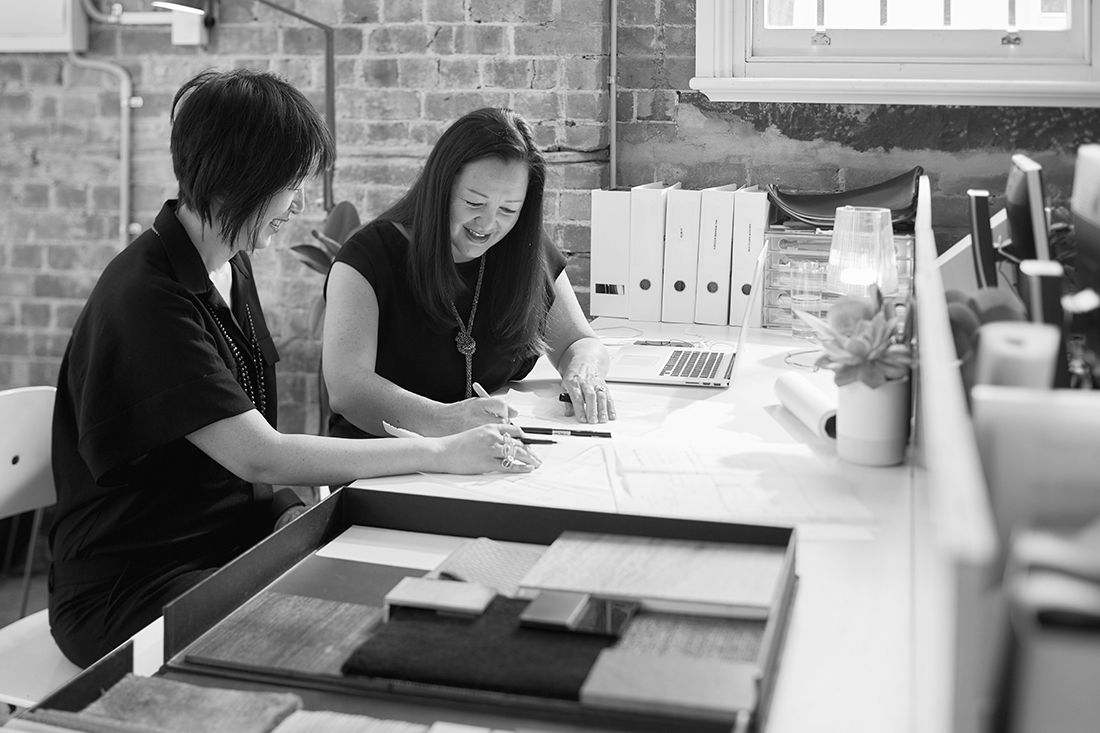 Two women working together at an office desk, looking at sample fabric or material swatches.