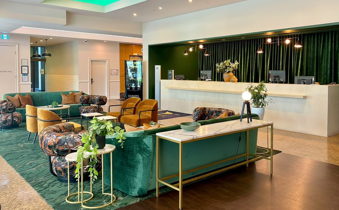 Modern hotel lobby with green and beige seating, a reception desk in the background, and decorative plants.