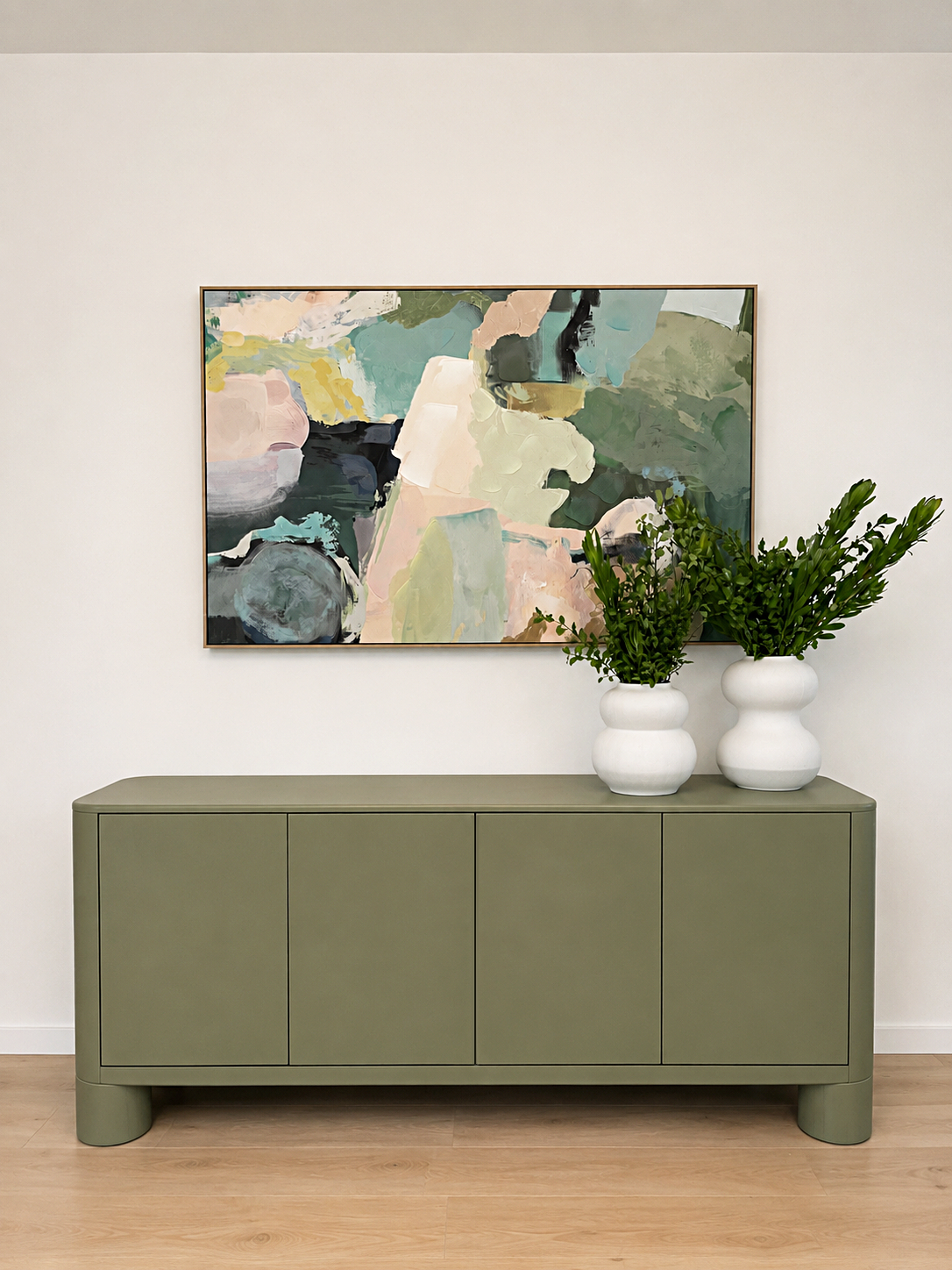 A light green sideboard with rounded legs, topped with two white vases containing green leafy plants, and a large abstract painting in pastel and dark colors on a white wall.