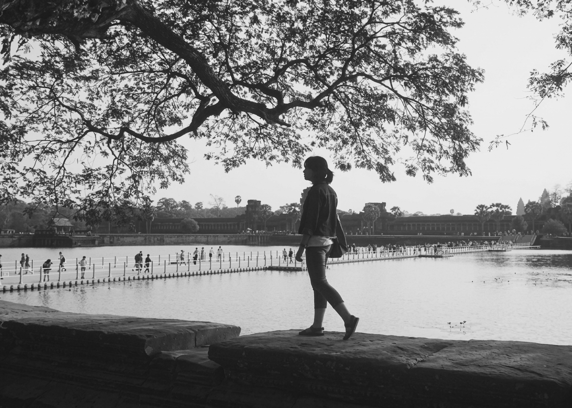 A girl walking near a lake with a large tree overhead, silhouettes with many people in the background, and historical structures across the water.