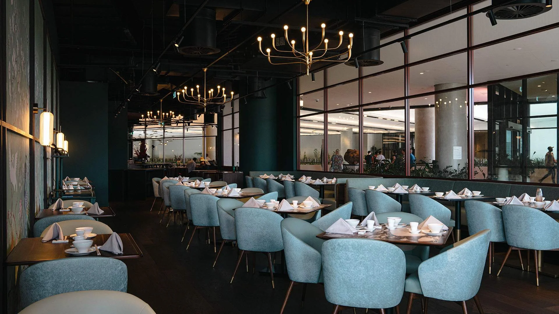 Interior of a modern restaurant with light blue chairs, dark wood tables set with white bowls, cups, and napkins, large windows, and elegant chandeliers.