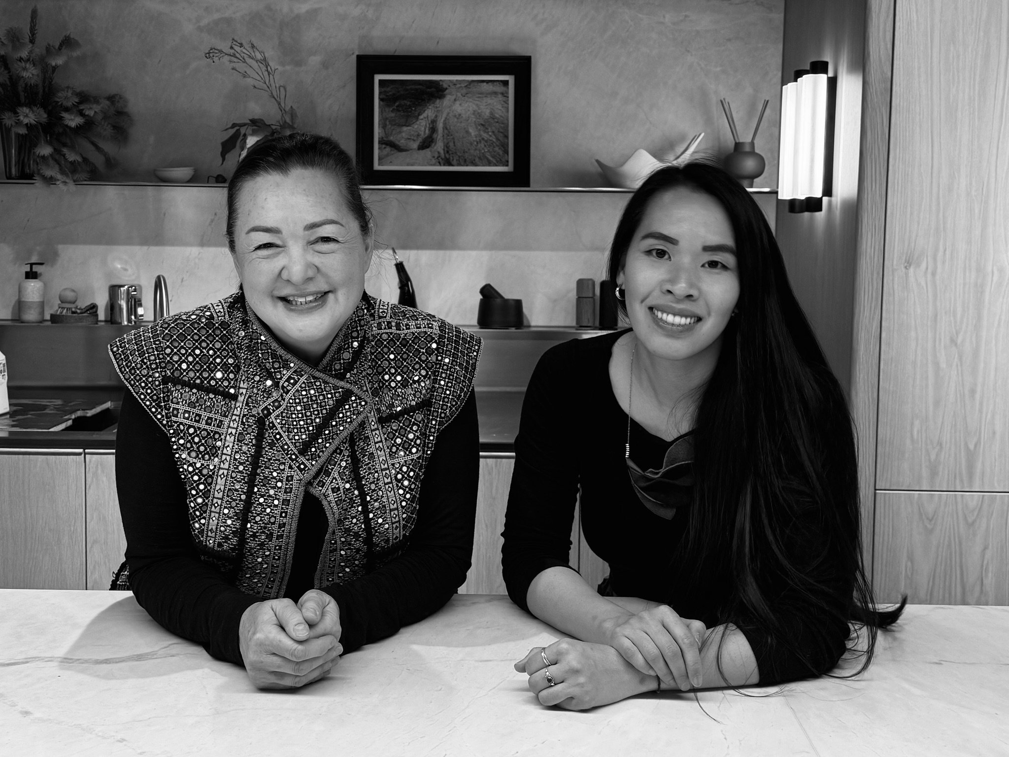Two women sitting at a table, smiling at the camera. The woman on the left has her hands clasped on the table and is wearing a patterned top. The woman on the right has long dark hair, is wearing a dark top, and has her hands on the table.