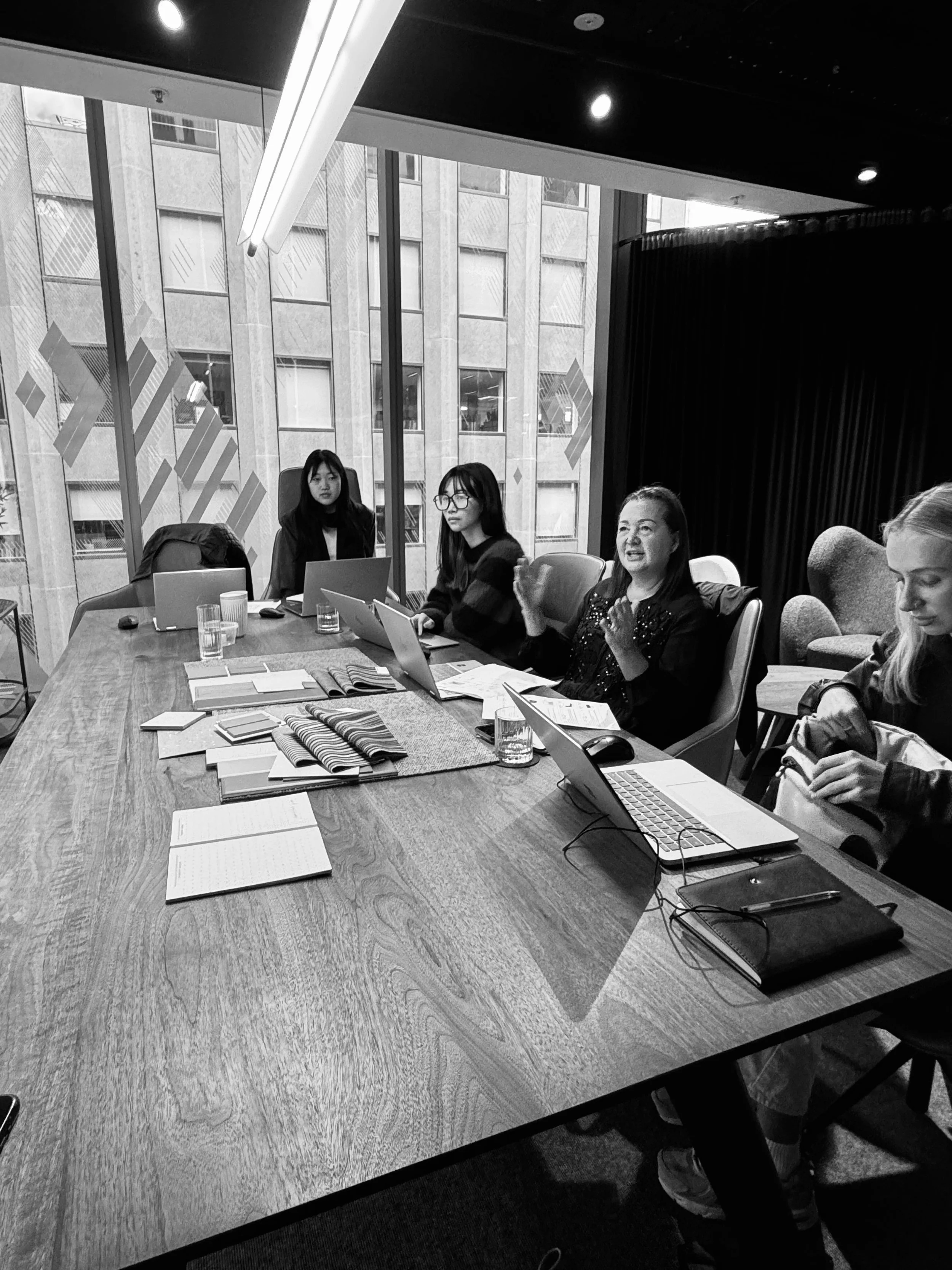 A black and white photo of a conference room with a large wooden table surrounded by people working on laptops. Four women are visible, one is speaking and clapping, others are listening or looking at their laptops. Large windows show tall buildings outside.
