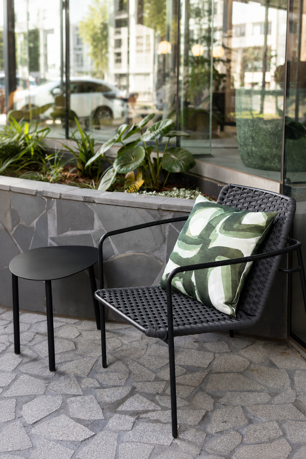 Outdoor seating area with a black woven chair with a green and white patterned cushion, small black side table, and a garden bed with green plants. Behind is a glass window reflecting cars and buildings.