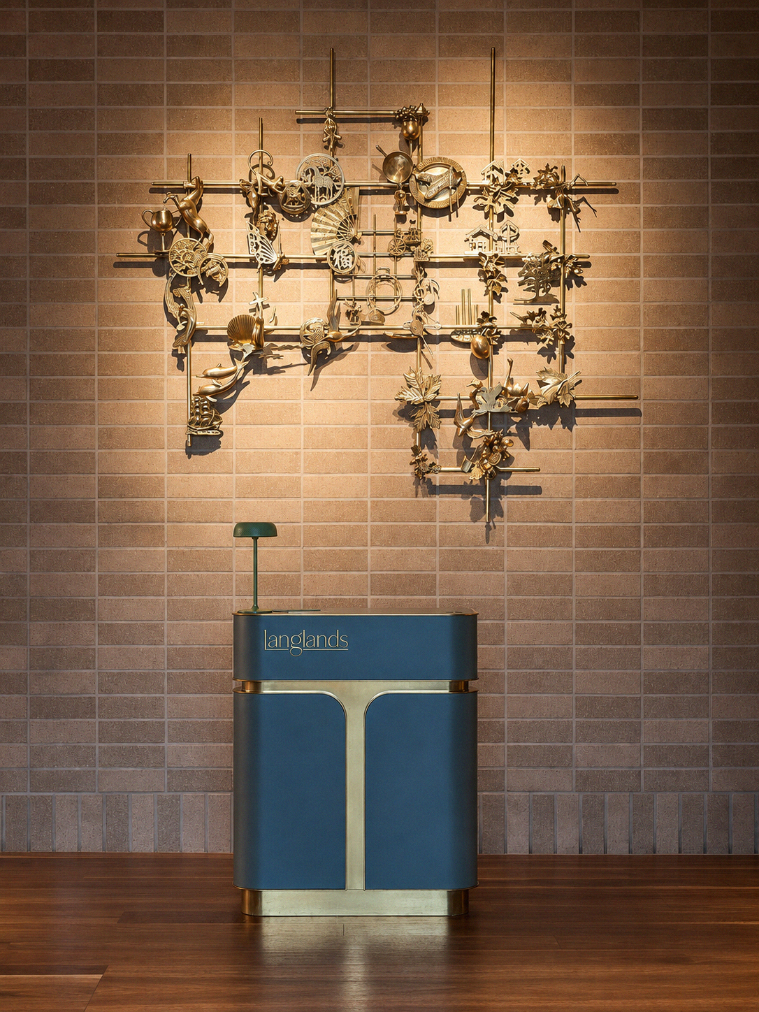 A modern interior with a decorative wall sculpture made of metallic objects shaped like various leaves, animals, and other designs, mounted on a brick wall. In front, there is a vintage-style blue cleaning cart with a green lamp on top, labeled 'Lang