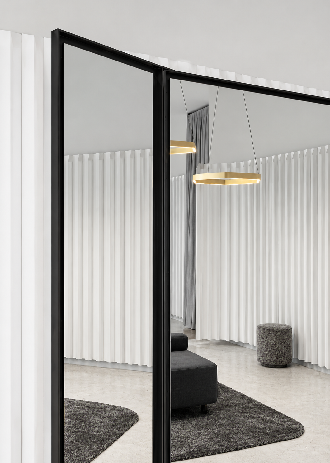 A large wall mirror in a modern interior space, reflecting a portion of a room with white, textured, vertical panel walls, a gray ottoman, and hanging gold geometric pendant lights.