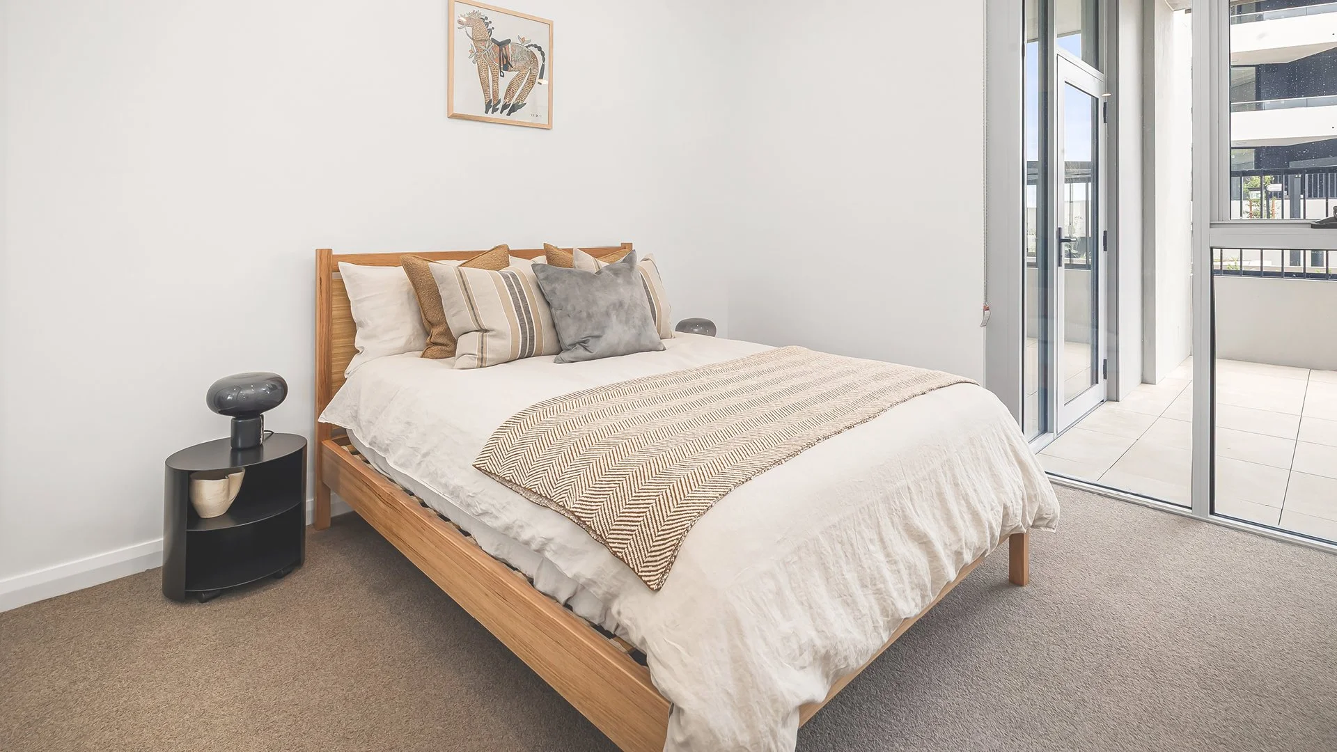 A bedroom with a wooden bed frame, cream and striped bedding, and multiple pillows. A black bedside table with a small lamp and decoration. Sliding glass doors leading to a balcony or patio with tiled flooring and black railing.
