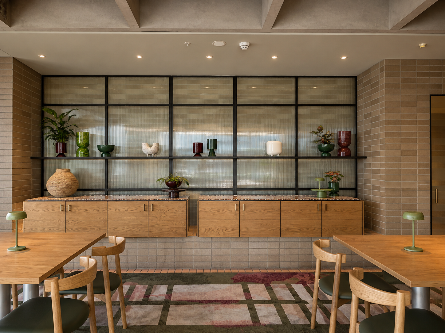 Interior of a modern dining or lounge area with wooden tables and chairs, decorative vases, and shelves with various plants and decorative objects against a brick wall and glass backdrop.