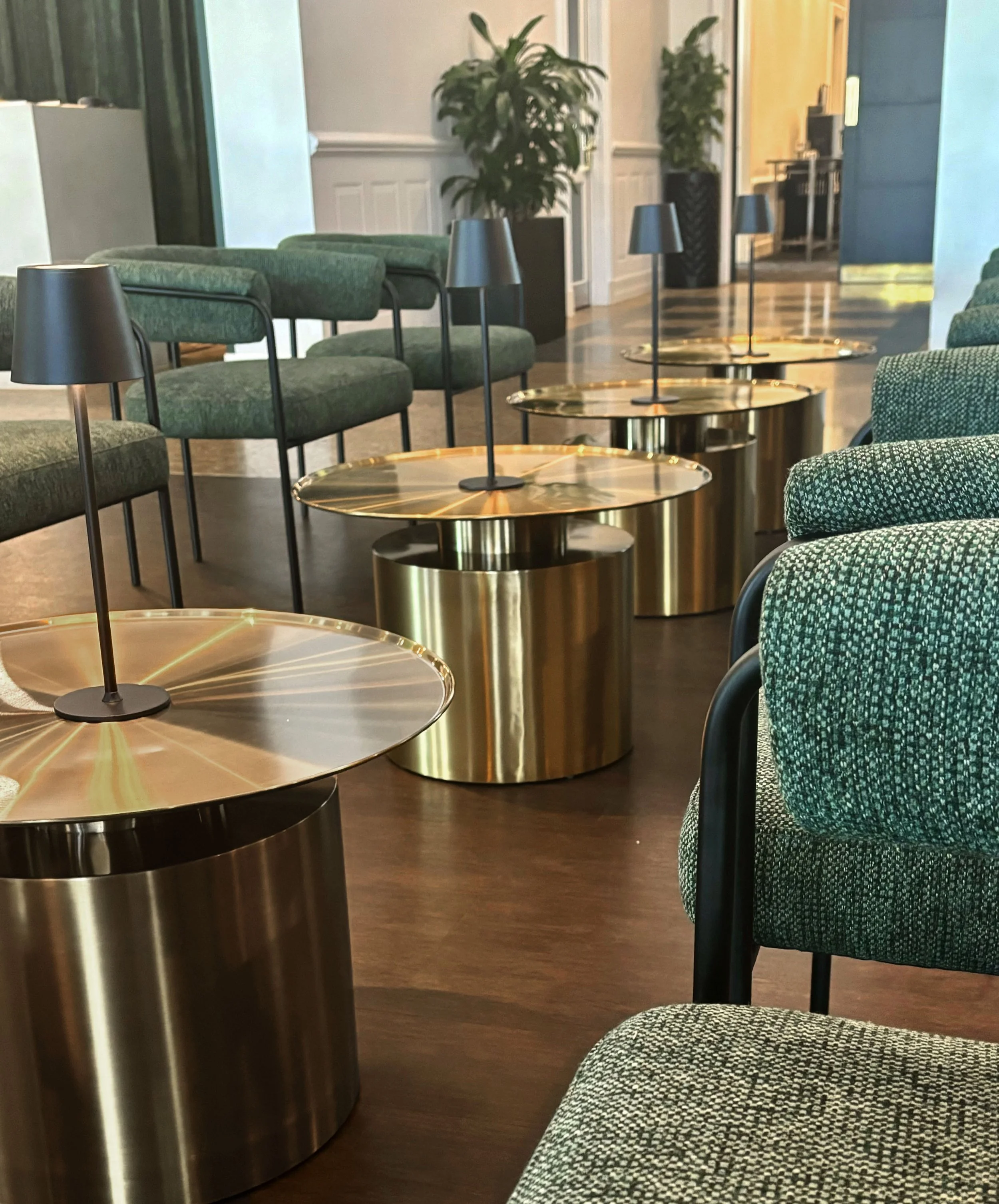 Modern interior with green upholstered chairs, round metallic side tables, and black lamps with cone-shaped shades along a wooden floor.