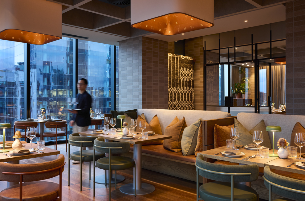 Interior of a modern upscale restaurant with floor-to-ceiling windows, wooden tables, cushioned chairs, and a beige sofa with pillows, set for dining with plates, glasses, and napkins, with a blurred server carrying dishes in the background.