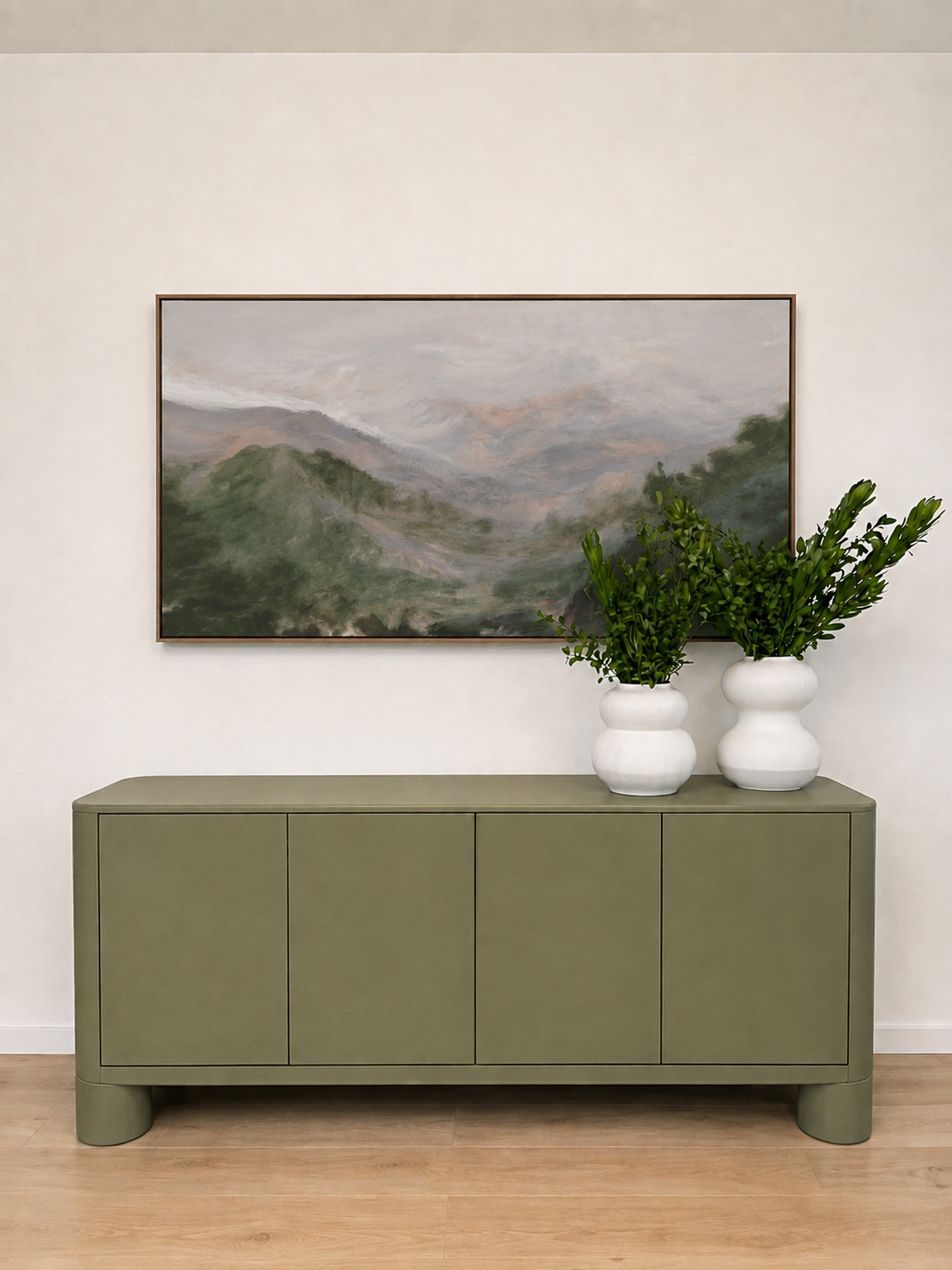A green sideboard with double doors and curved legs, two white vases with green leafy plants, a mountain landscape painting, and a white wall background with wooden flooring.