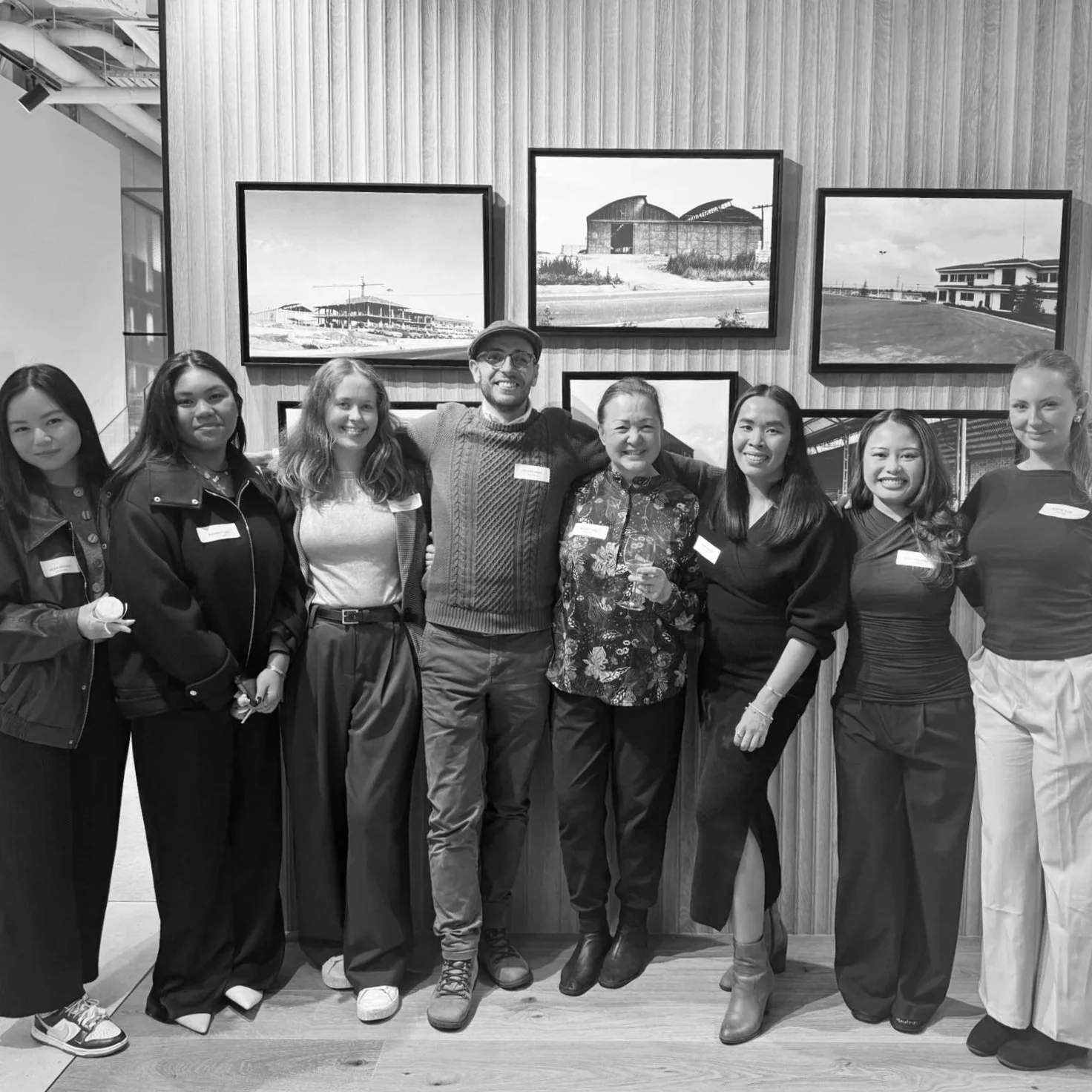 Group of eight diverse women and one man standing close together, smiling, in front of a wall with framed photos.