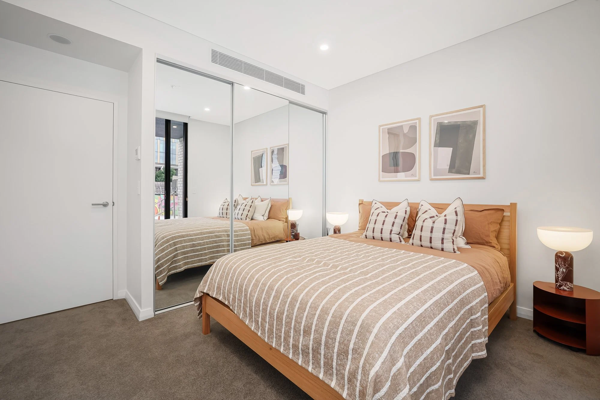 A bedroom with a double bed, striped bedding, and multiple pillows, featuring a large mirror closet, artwork on the wall, and a nightstand with a lamp.