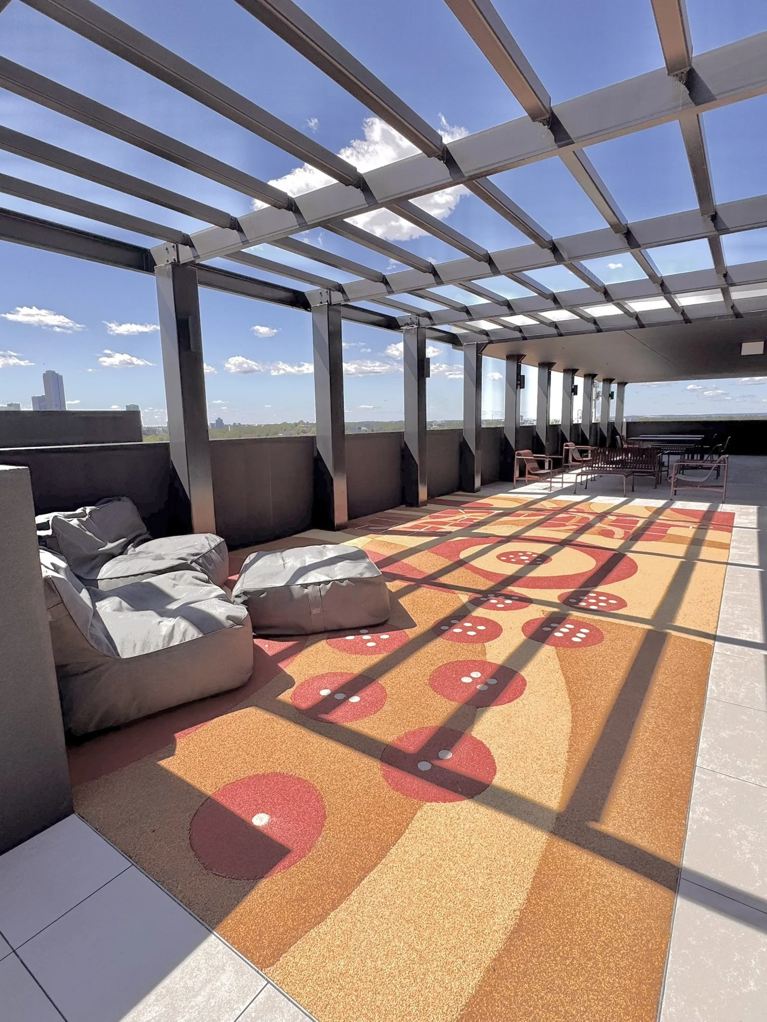 Rooftop terrace with bean bag chairs, a colorful patterned carpet, and outdoor seating, under a metal pergola with shadows cast by the structure on a sunny day.