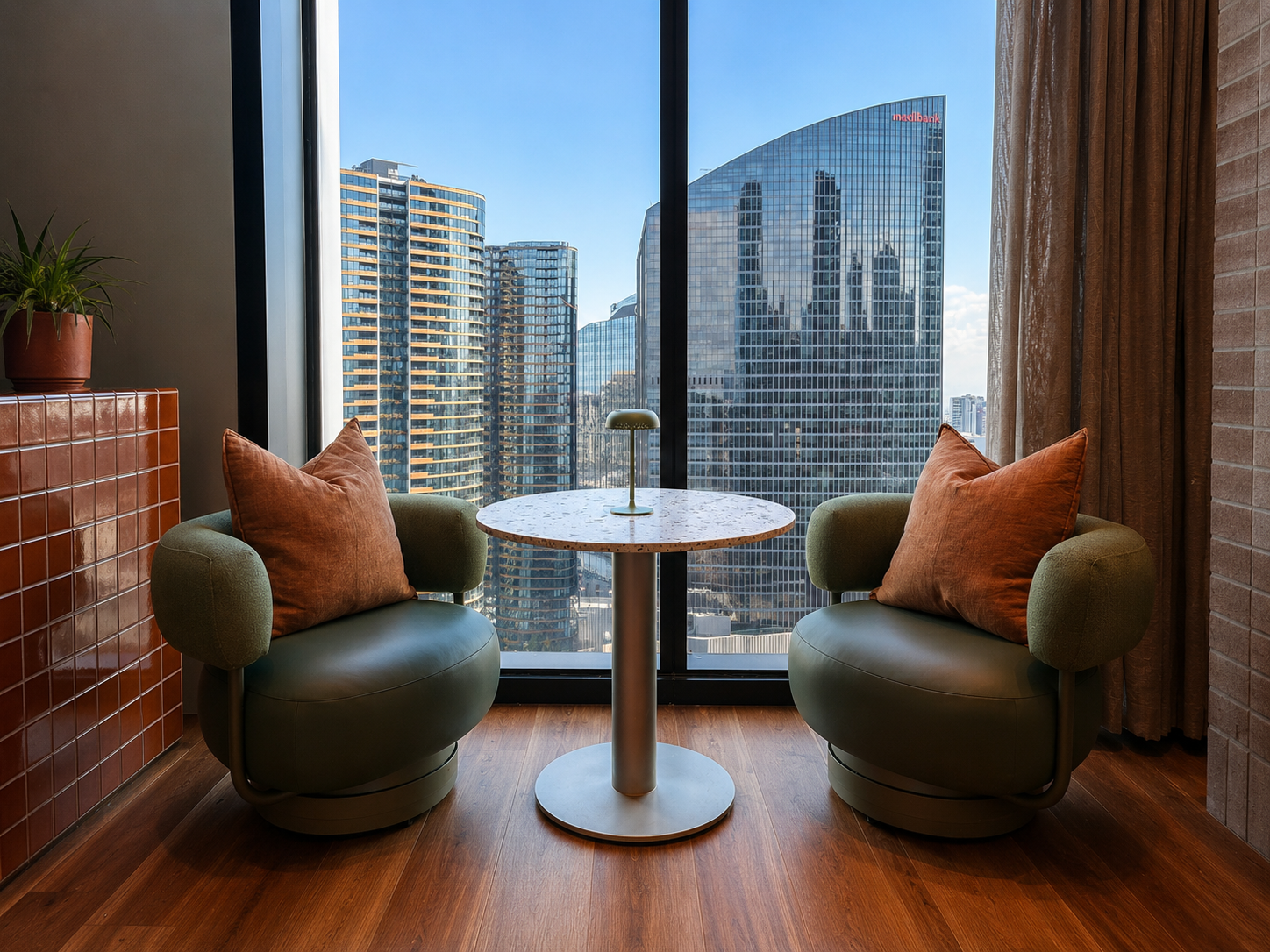 Interior view of a high-rise apartment with two green armchairs with brown pillows on a wooden floor, facing a small round marble table with a modern lamp, large floor-to-ceiling window with city skyscrapers and blue sky outside, beige curtains on th