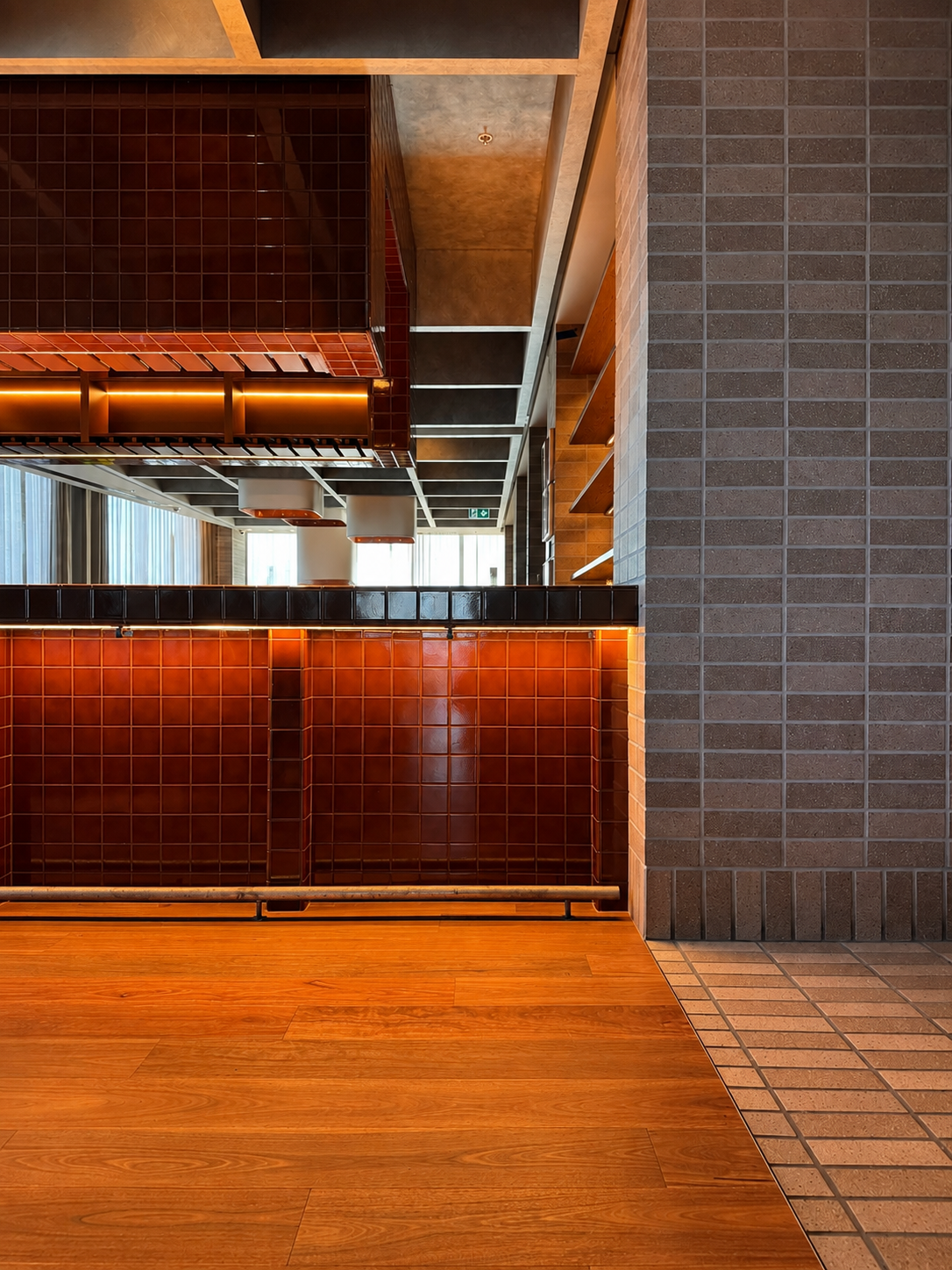 Indoor restaurant or cafe interior featuring a wooden floor, dark red tilework, and brick walls with open shelving. Light filters in through large windows.