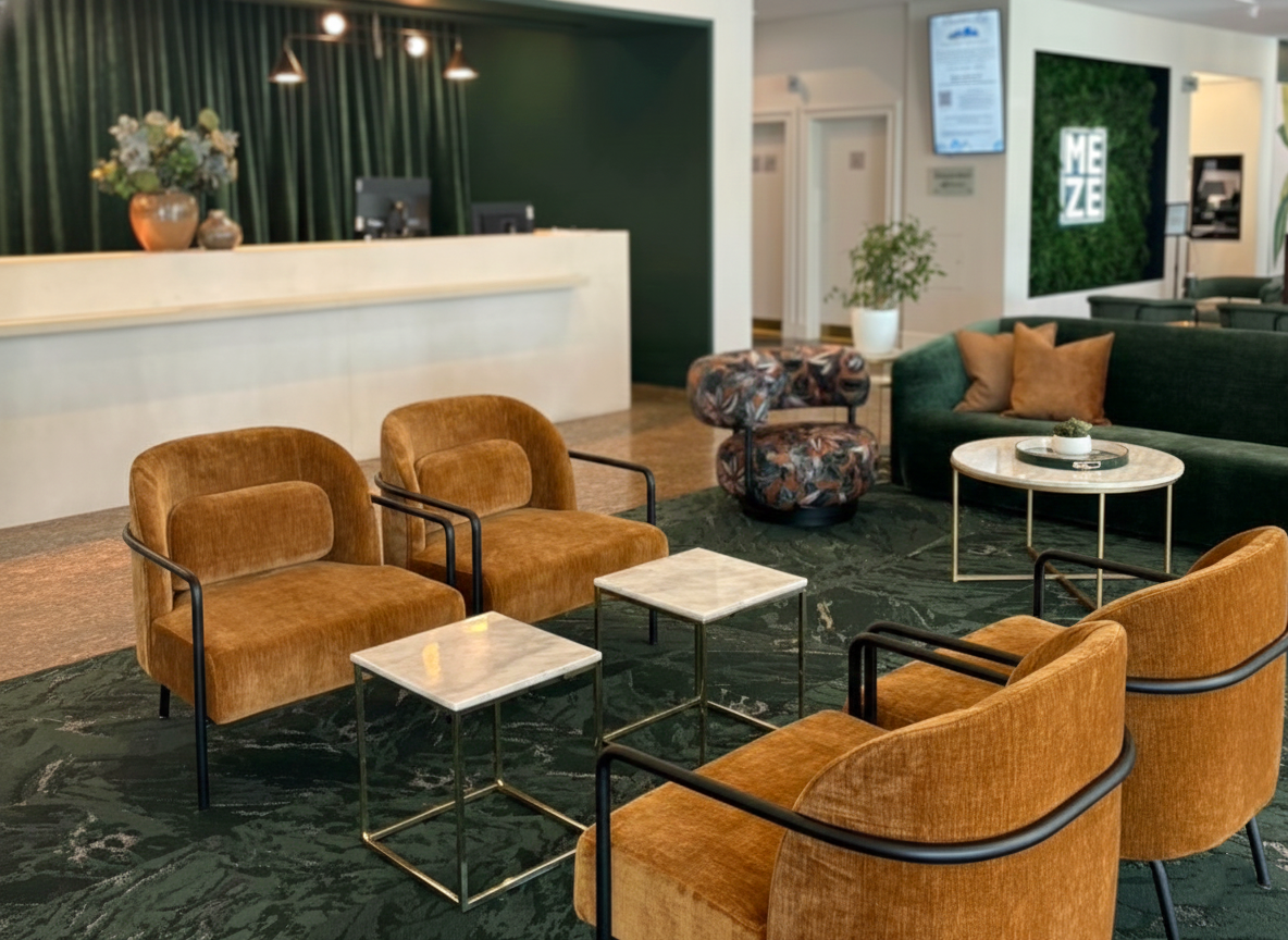 Lobby with a front desk, green curtains, a flower arrangement, seating area with brown and green upholstered chairs and a small round table, and wall decor including a sign with "MEZE."