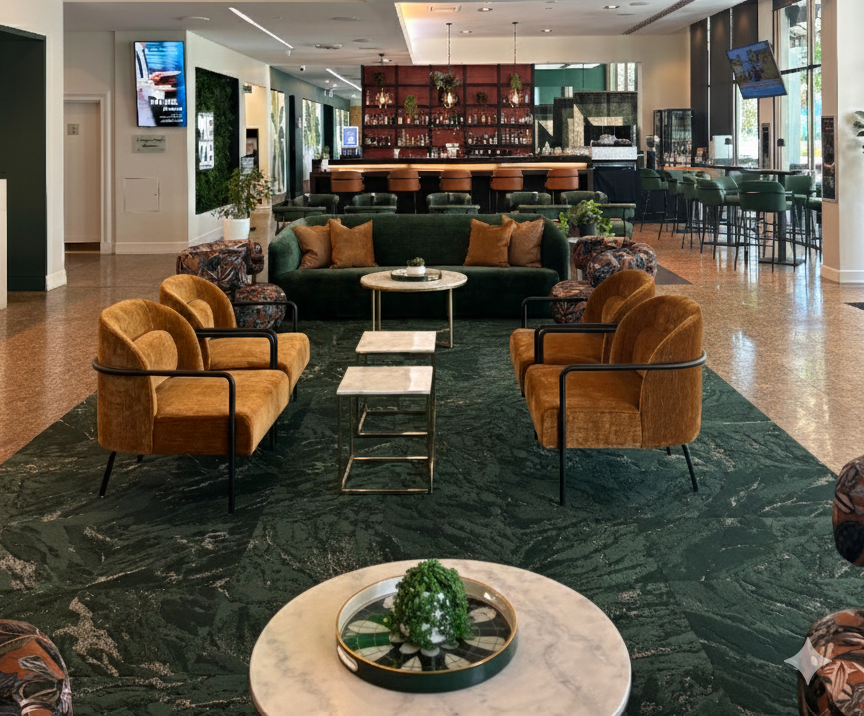 Elegant hotel lobby with green marble flooring, a central seating area with a dark green sofa and tan armchairs, and a bar area with green chairs and a red accent wall in the background.