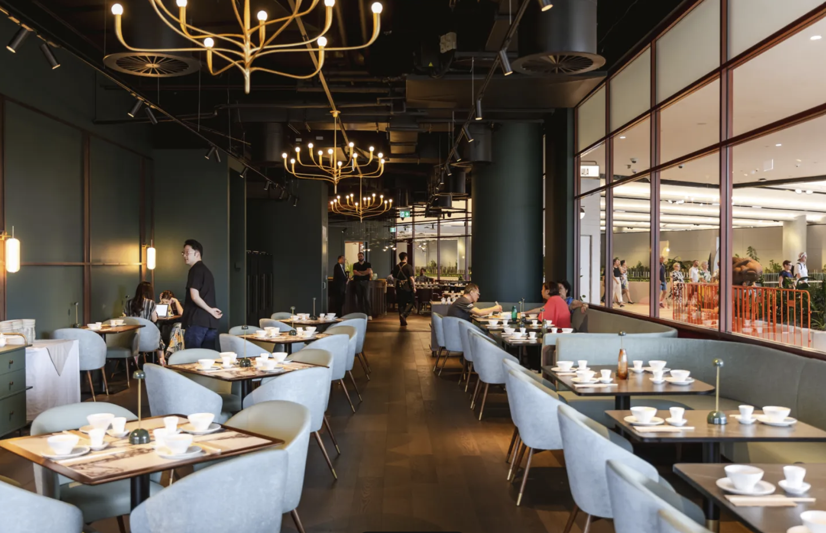 Interior view of a modern restaurant with tables set for dining. There are light gray chairs, dark wooden tables, and chandeliers hanging from a dark ceiling. Large windows show people walking outside.