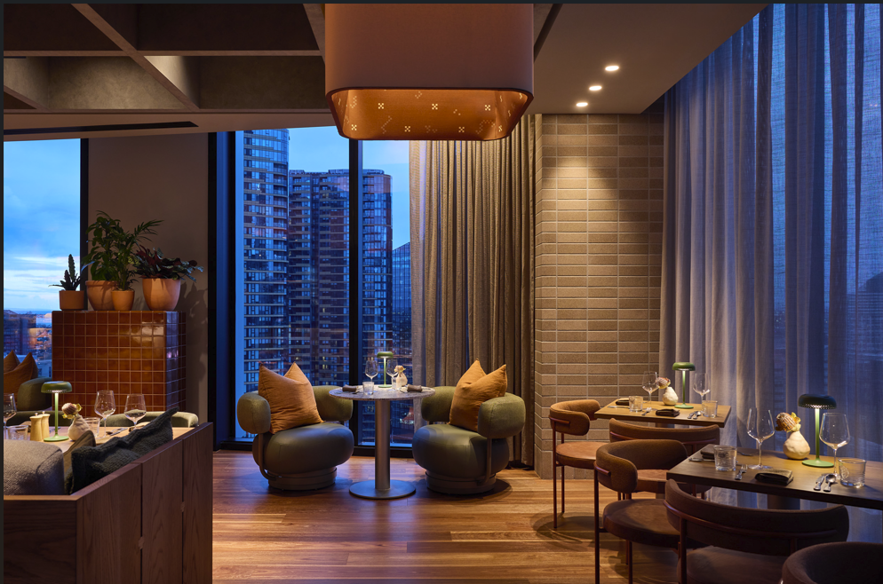 Elegant restaurant with large windows, city skyline view, wooden flooring, modern chairs with cushions, small tables set with glassware and decor, and potted plants, lit with warm lighting.