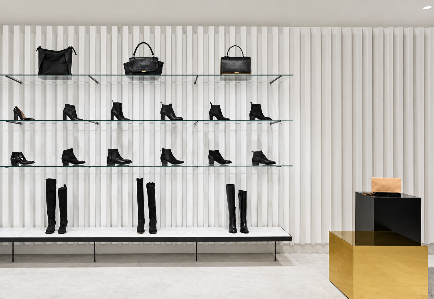 Display of black handbags, boots, and shoes on glass shelves against a textured white wall, with a black and gold pedestal furniture piece holding a beige clutch purse.