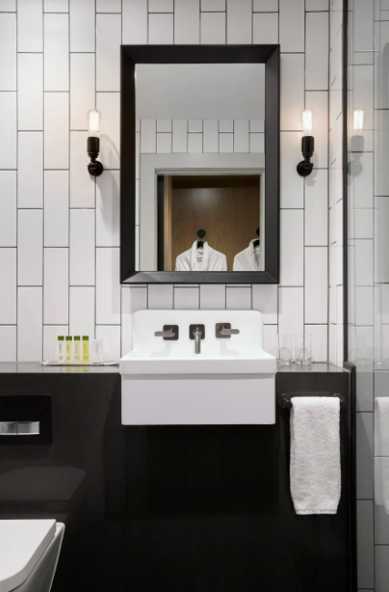 Modern bathroom with a white sink, black framing mirror, wall-mounted lights, and white tiled walls.