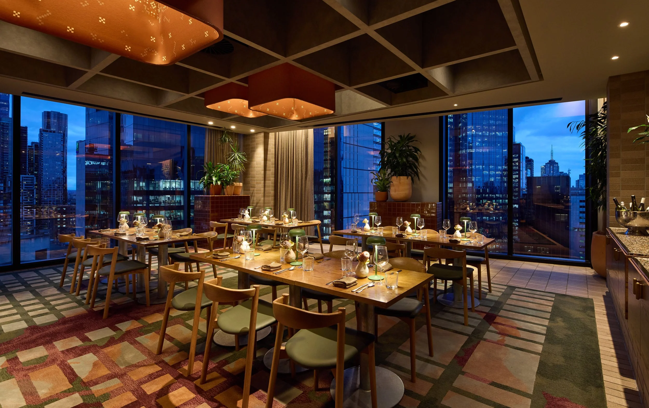 Interior of a modern restaurant with wooden tables and chairs, large windows showing a city skyline at dusk, decorative plants, and ceiling lighting fixtures.