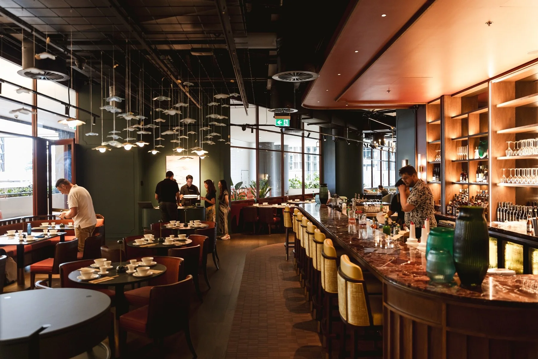 Interior of a modern restaurant with a bar counter, ceiling decorations resembling fish, and large windows letting in natural light. Several people are preparing and setting up, and the restaurant appears to be ready for guests.