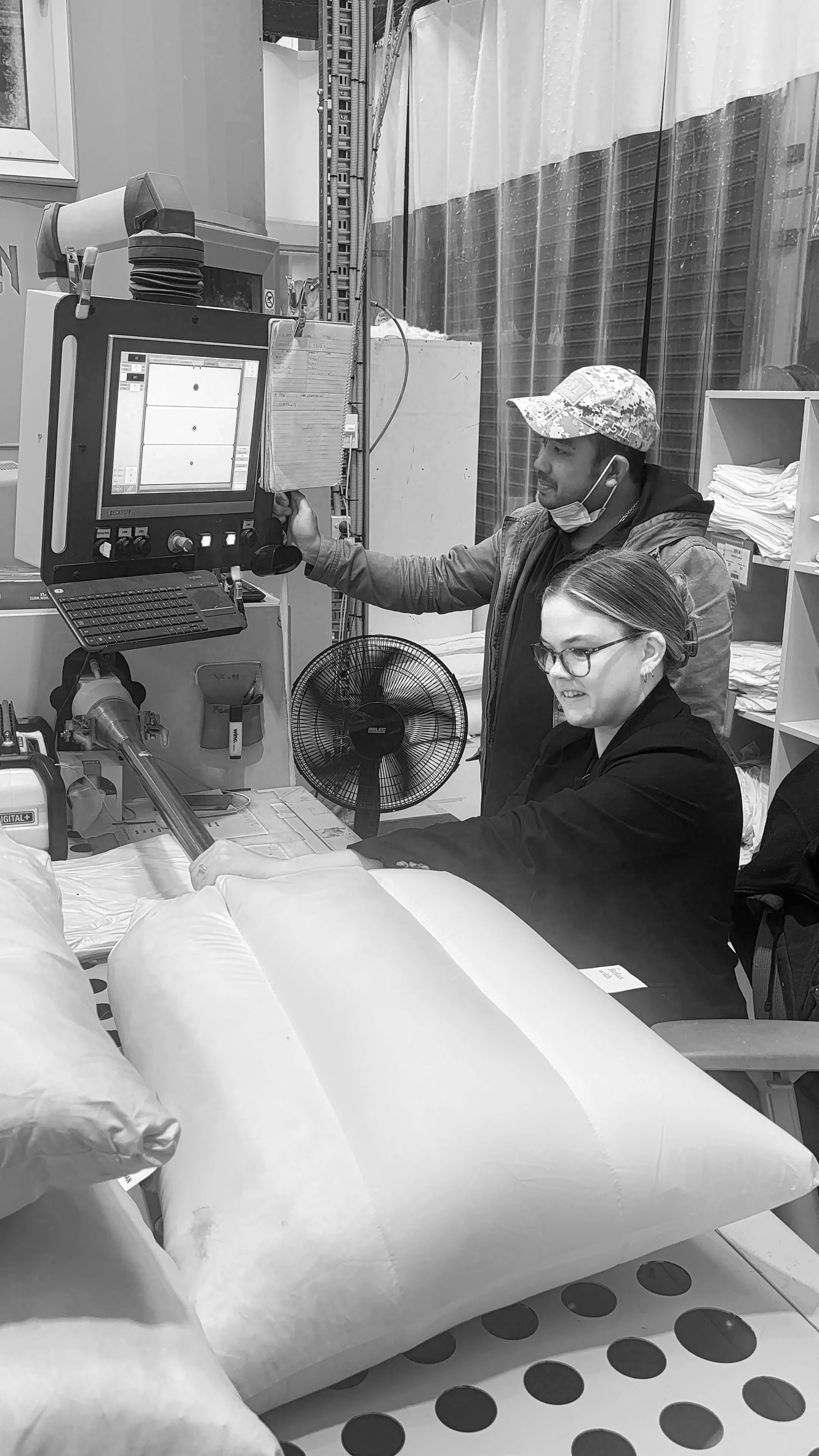 A man and a woman appear to be working in a medical or laboratory setting. The man is standing and looking at a monitor, while the woman is sitting and working with some equipment, with a large package or pillow in the foreground. Both are wearing masks, and the woman has glasses and has her hair tied back.
