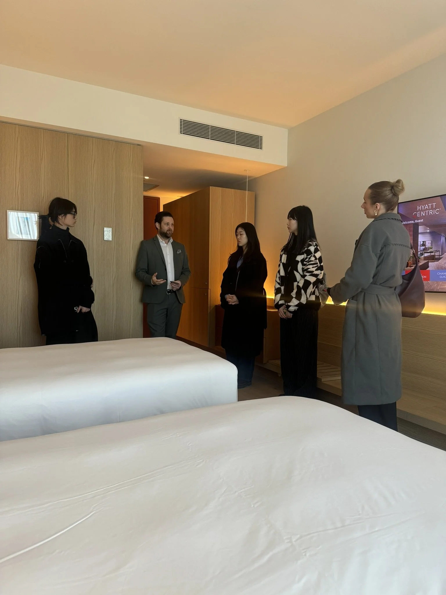 Group of five people standing in a hotel room, having a conversation; two women, one man, and two more women, all dressed in business or casual attire.
